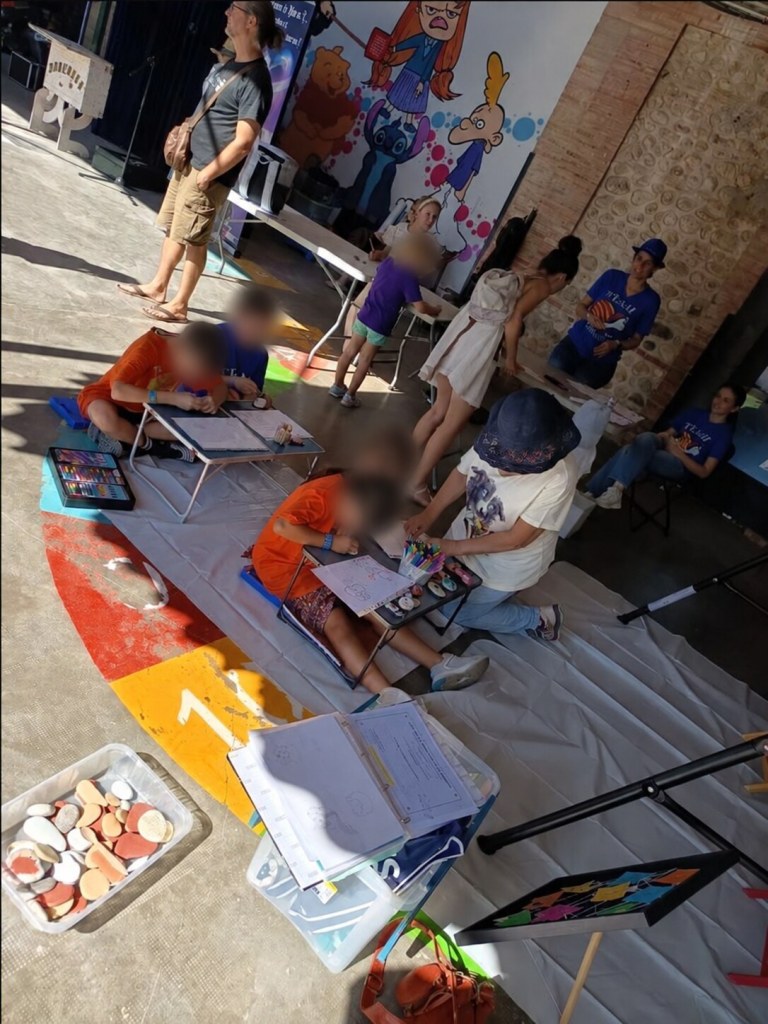 Plusieurs enfants dessinent et décorent des galets sur le stand d’ASCaMaC, leurs visages sont floutés.
