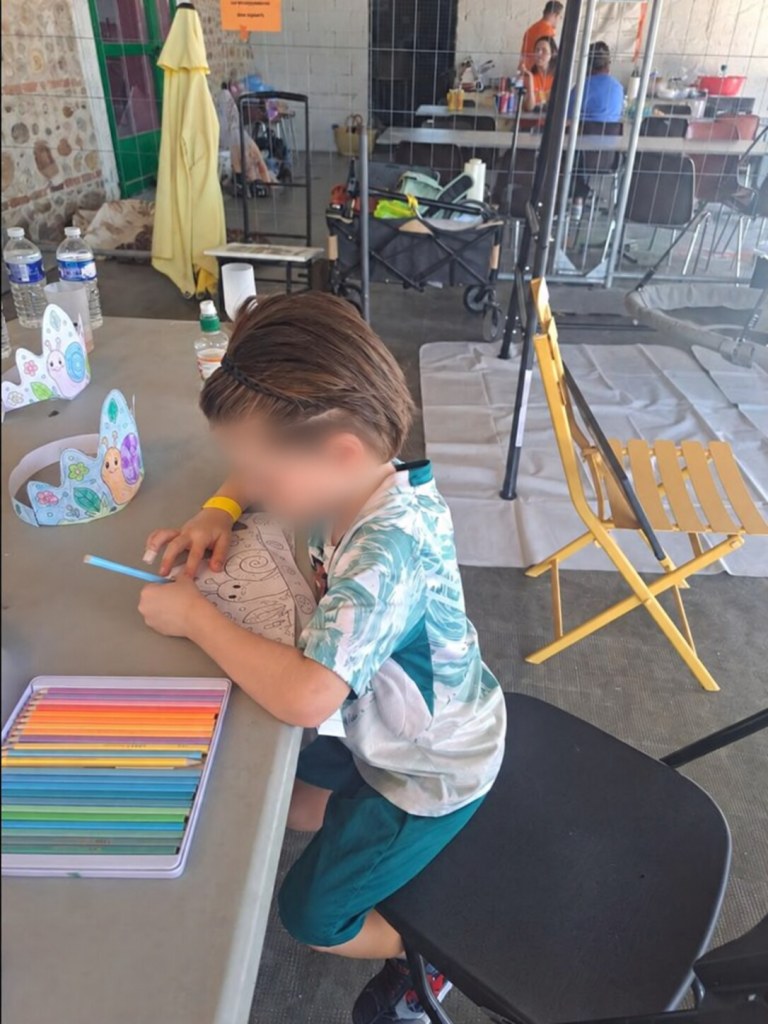 Un enfant dessine à une table avec une boîte de crayons de couleur, son visage est flouté.