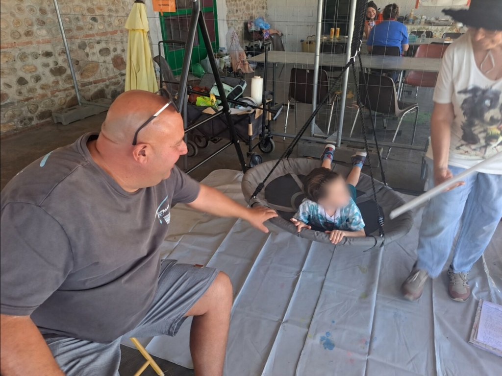 Un enfant installé sur une balançoire circulaire participe à l’activité, son visage est flouté, entouré de deux adultes.